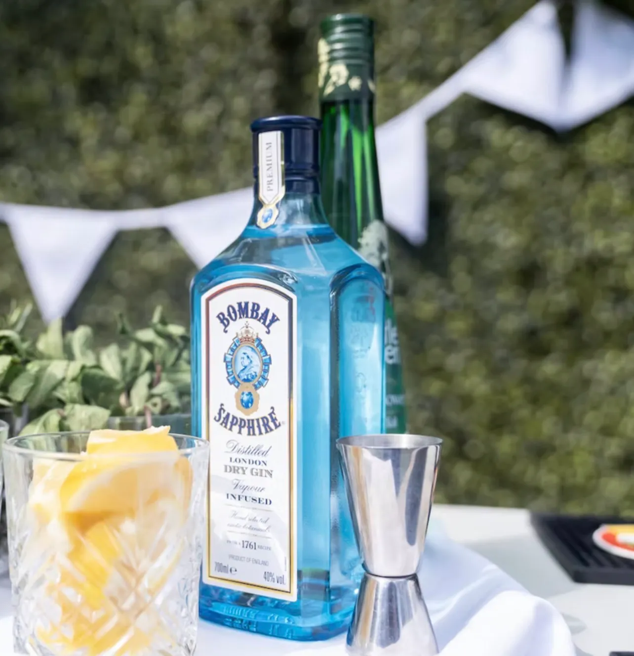 A bottle of gin and a bottle of elderflower on a linen-covered bar, a cut-glass tumbler of lemon wedges and a steel jigger beside them and bunting strung behind