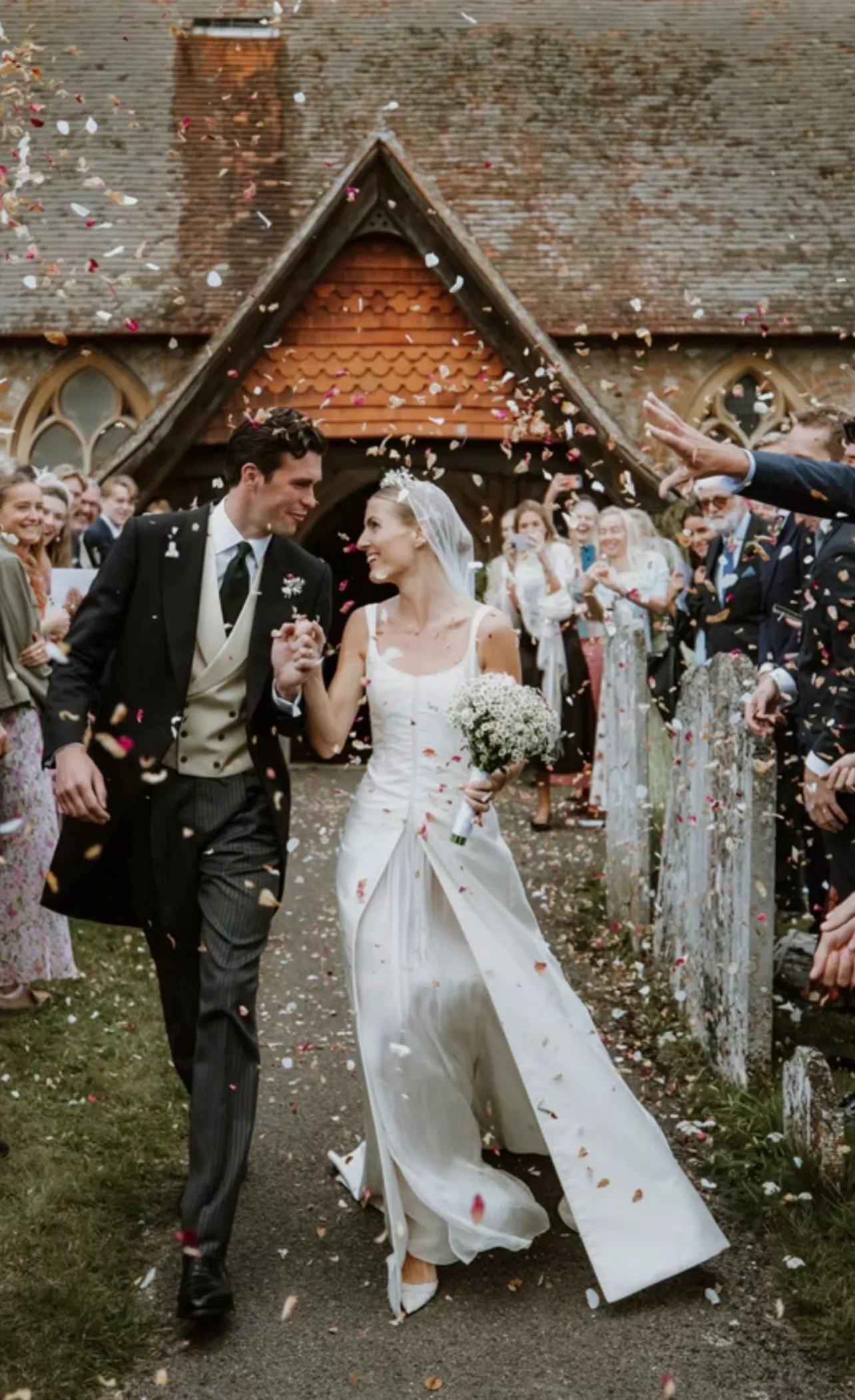 A bride and groom walking hand in hand from a church porch through confetti thrown by guests lining the path on both sides