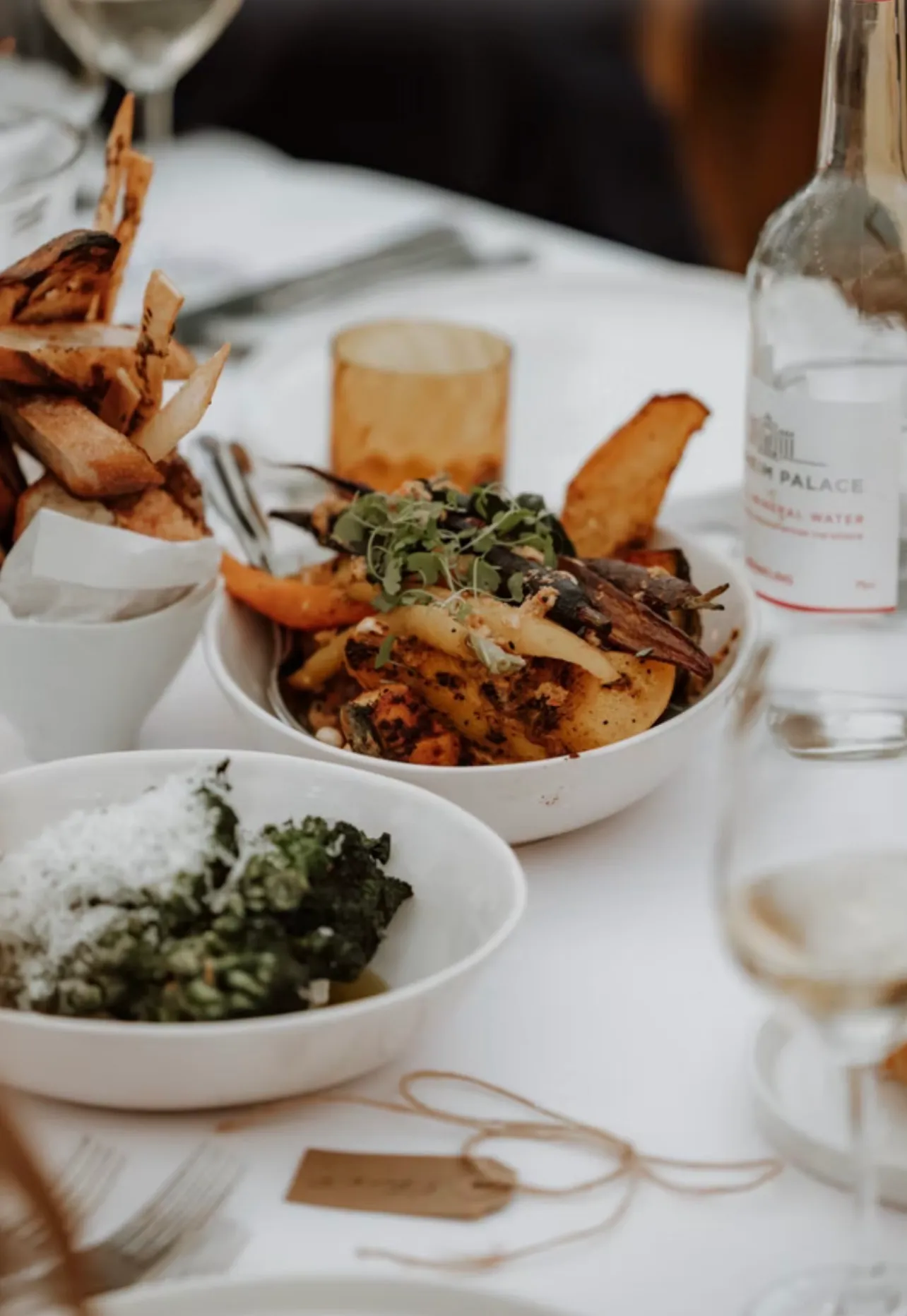 Sharing bowls of roast vegetables, greens and chips set on a white cloth between glasses and a bottle of water