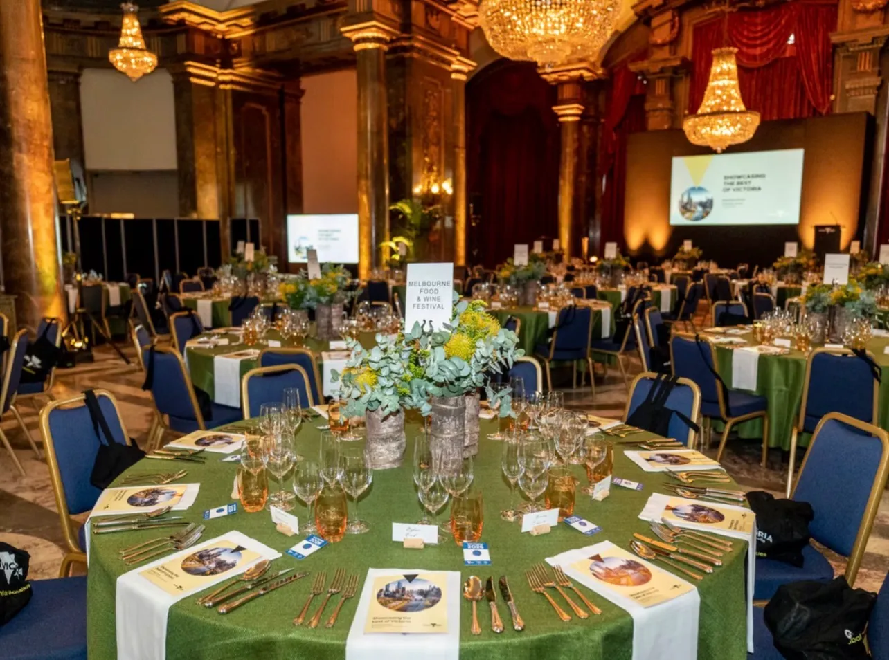 Round tables dressed in green under chandeliers in a gilded hall, places laid with menus and glassware and presentation screens behind