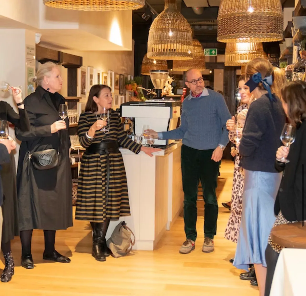 Guests standing with wine glasses around a counter while a man speaks to them, woven pendant lights hanging above