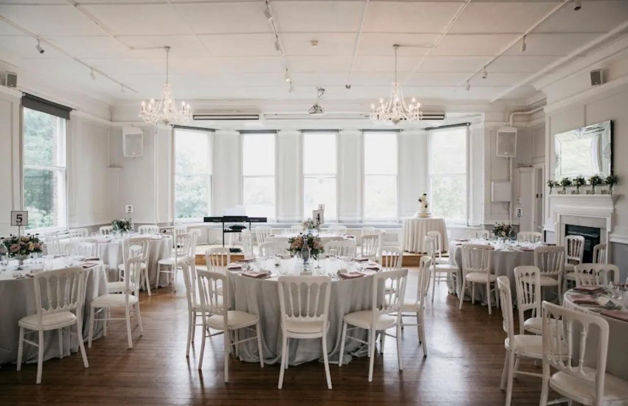 Round tables laid for a wedding breakfast under chandeliers, chairs set out across a panelled room with bay windows